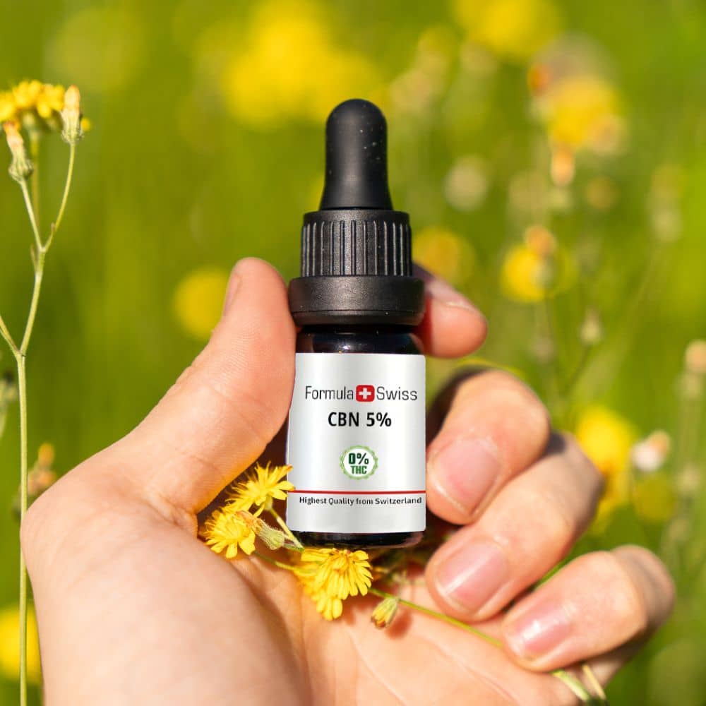 Hånd, der holder en flaske Formula Swiss CBN olie 5 % mellem gule blomster